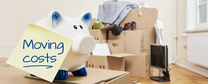 On a moving box there is a piggy bank on which a yellow piece of paper sticks. The note is in English language. On the note is moving coats. In the background is a stack of packed moving boxes