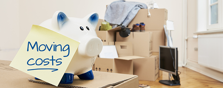 On a moving box there is a piggy bank on which a yellow piece of paper sticks. The note is in English language. On the note is moving coats. In the background is a stack of packed moving boxes