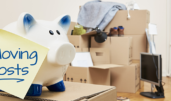 On a moving box there is a piggy bank on which a yellow piece of paper sticks. The note is in English language. On the note is moving coats. In the background is a stack of packed moving boxes